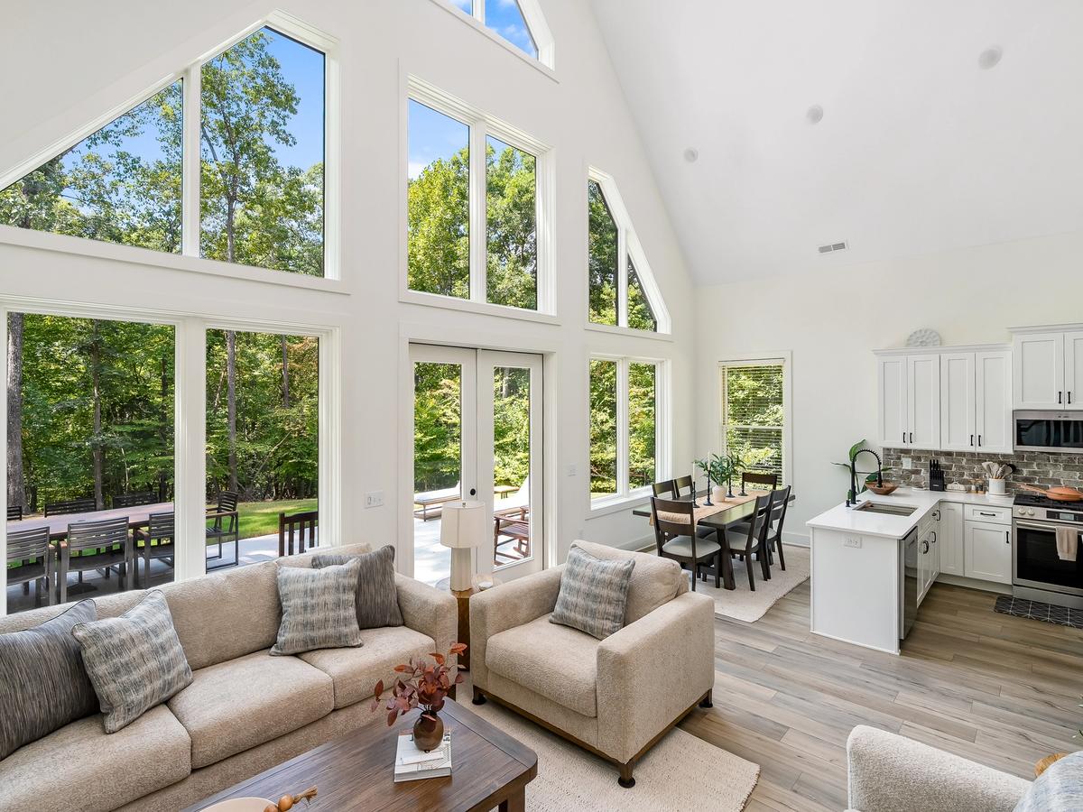 Modern open-concept living room and kitchen with vaulted ceilings, large floor-to-ceiling windows, neutral-toned sofa and chairs, dining area, and white cabinetry with stainless steel appliances.