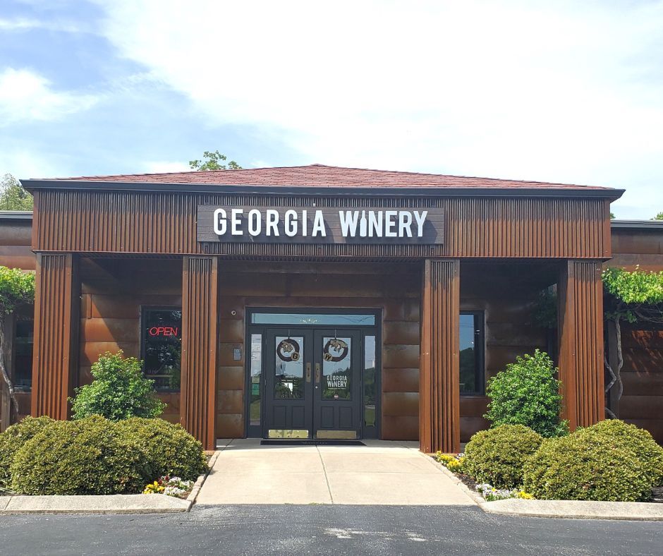 This photo shows the front entrance of Georgia Winery, with a clean walkway leading to double doors under a bold sign. The building has warm brown tones, giving it a cozy and welcoming feel. Bright greenery around the entrance adds a touch of charm.