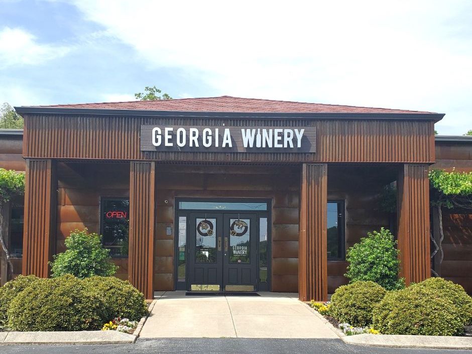 This photo shows the front entrance of Georgia Winery, with a clean walkway leading to double doors under a bold sign. The building has warm brown tones, giving it a cozy and welcoming feel. Bright greenery around the entrance adds a touch of charm.