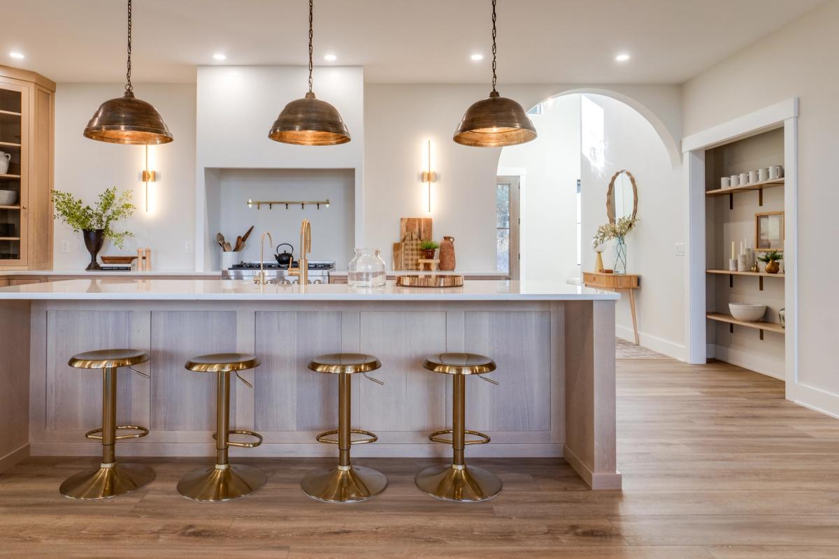 A warm modern kitchen with brass stools, pendant lights, and a bright white island countertop. The natural wood floors and open shelving give this space a cozy, high-end beach house feel.
