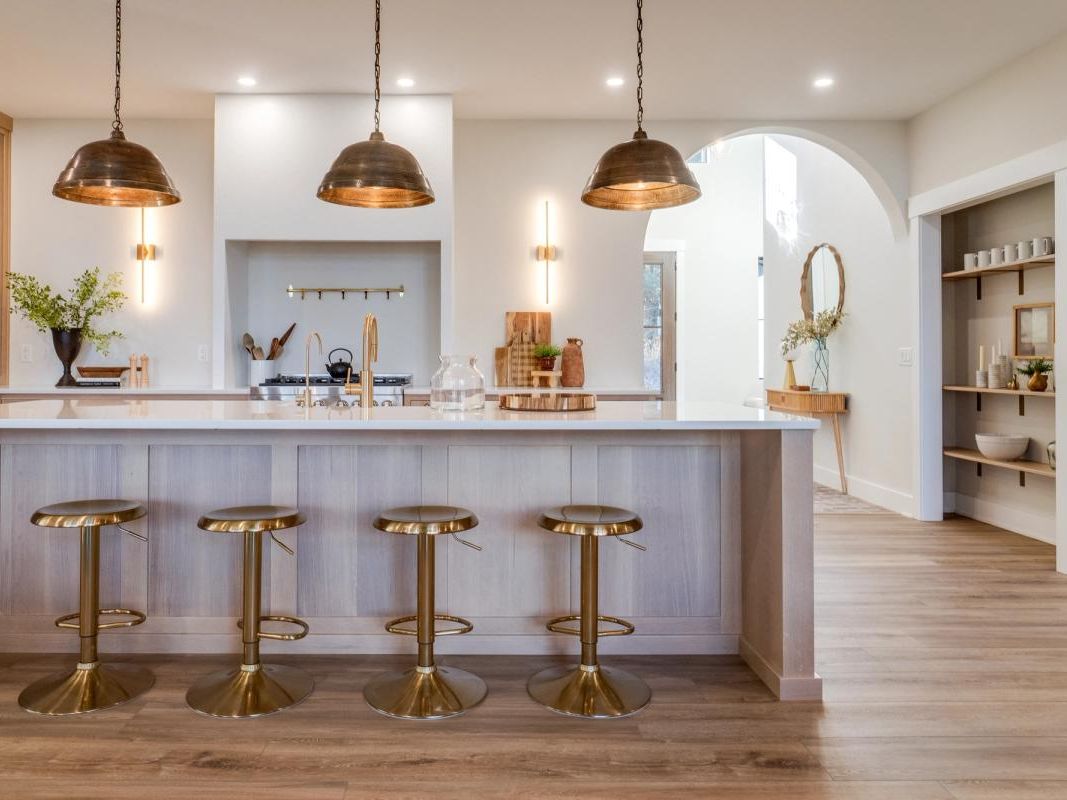 A warm modern kitchen with brass stools, pendant lights, and a bright white island countertop. The natural wood floors and open shelving give this space a cozy, high-end beach house feel.