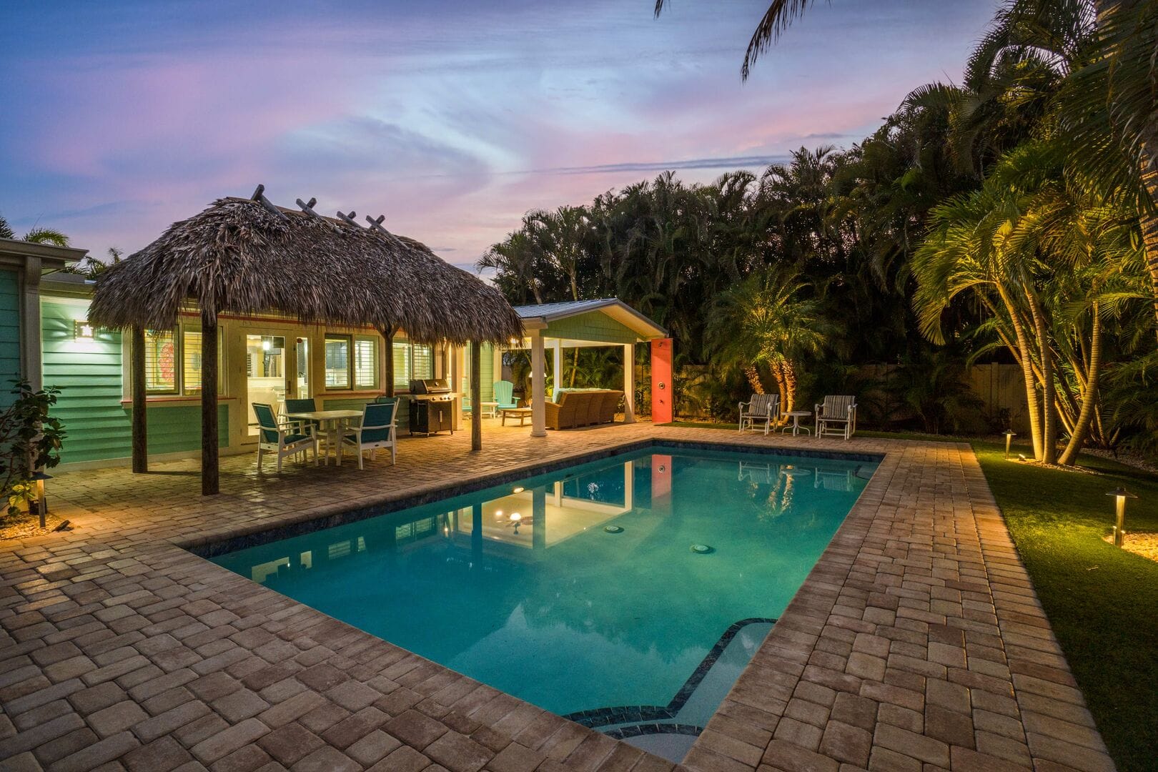 At dusk, a private tropical backyard glows with warm lighting around a tranquil swimming pool, featuring a thatched tiki-style pavilion, covered patio seating, and lush palm-lined surroundings under a pastel evening sky.