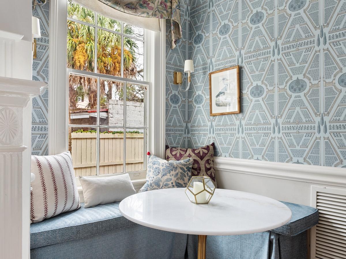 A cozy breakfast nook with a round marble table and upholstered bench seating is set against patterned blue wallpaper, featuring plush pillows, a framed bird print, and a large window overlooking a palm-lined yard.