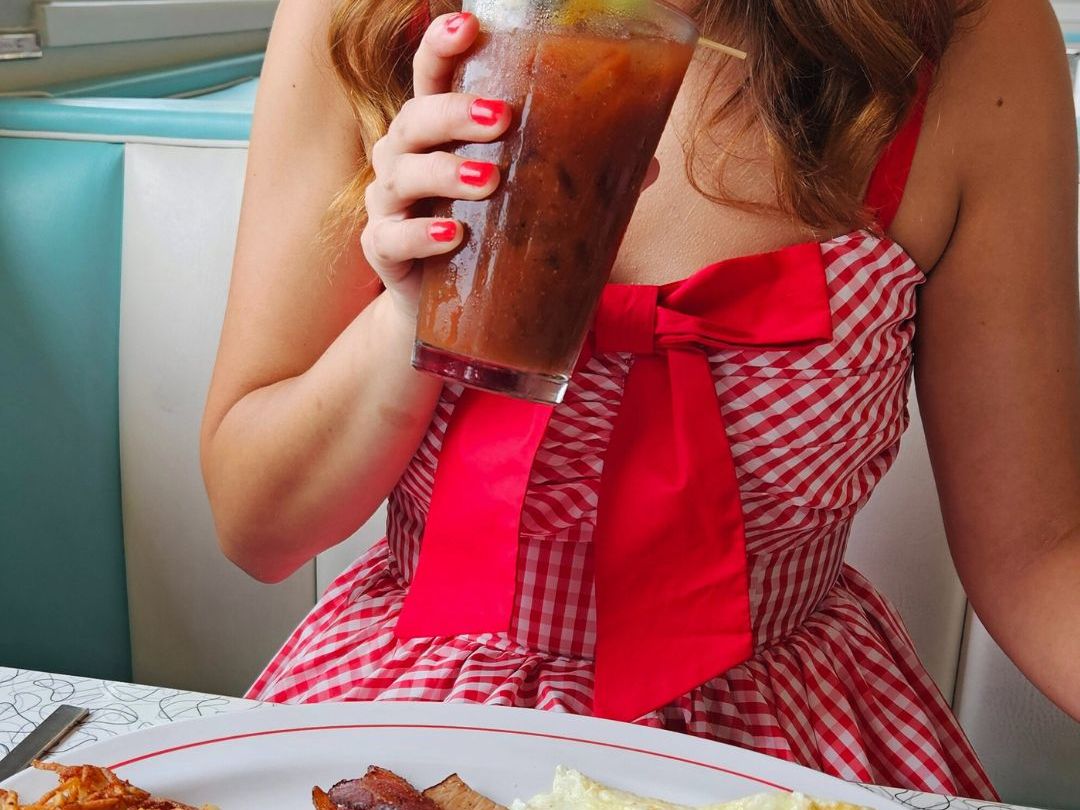 Retro diner breakfast with eggs, bacon, hash browns, and a Bloody Mary, enjoyed by a woman in a red gingham dress. A vintage-style brunch made for Instagram.