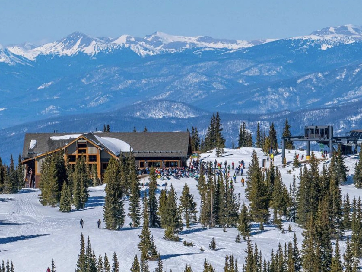 Snow-covered ski lodge surrounded by pine trees and skiers, set against a stunning backdrop of layered mountain ranges under a clear blue sky.