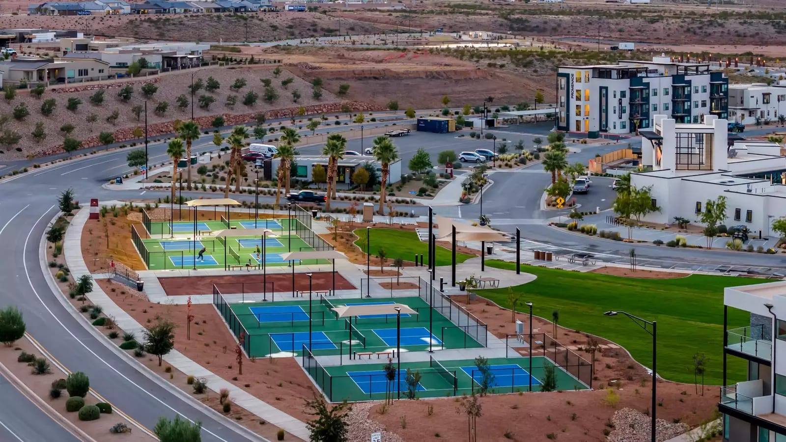 An aerial view of a modern outdoor pickleball and sports complex in St. George, Utah, features multiple blue and green courts set against the region's characteristic red rock desert landscape. Palm trees, manicured green space, and contemporary mixed-use buildings surround the facility, with players visible on the courts below.