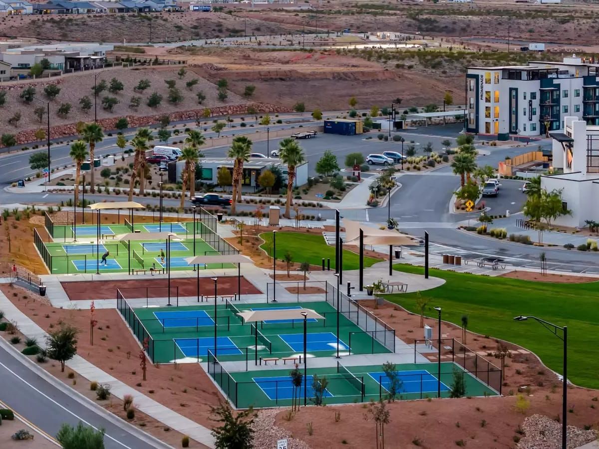 An aerial view of a modern outdoor pickleball and sports complex in St. George, Utah, features multiple blue and green courts set against the region's characteristic red rock desert landscape. Palm trees, manicured green space, and contemporary mixed-use buildings surround the facility, with players visible on the courts below.