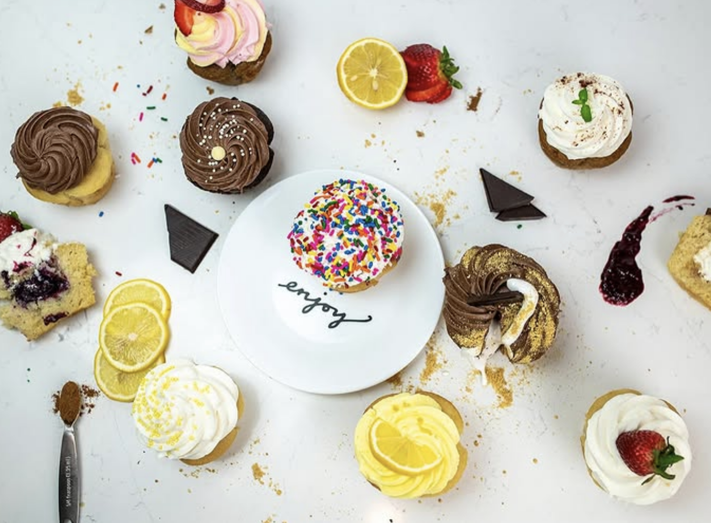 An assortment of colorful cupcakes with various frostings and toppings, including chocolate, lemon, berries, and sprinkles, is arranged around a white plate that says "enjoy" on a white surface.