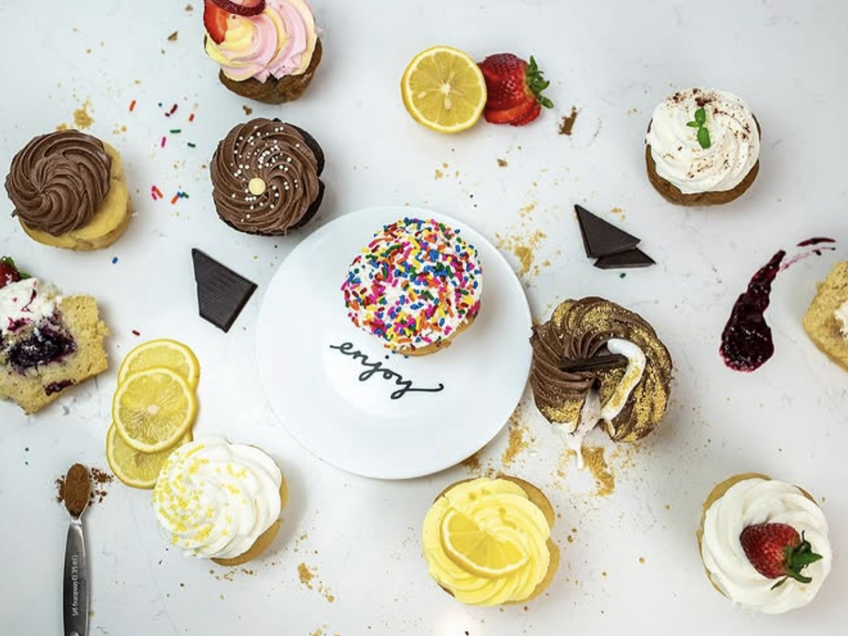 An assortment of colorful cupcakes with various frostings and toppings, including chocolate, lemon, berries, and sprinkles, is arranged around a white plate that says "enjoy" on a white surface.