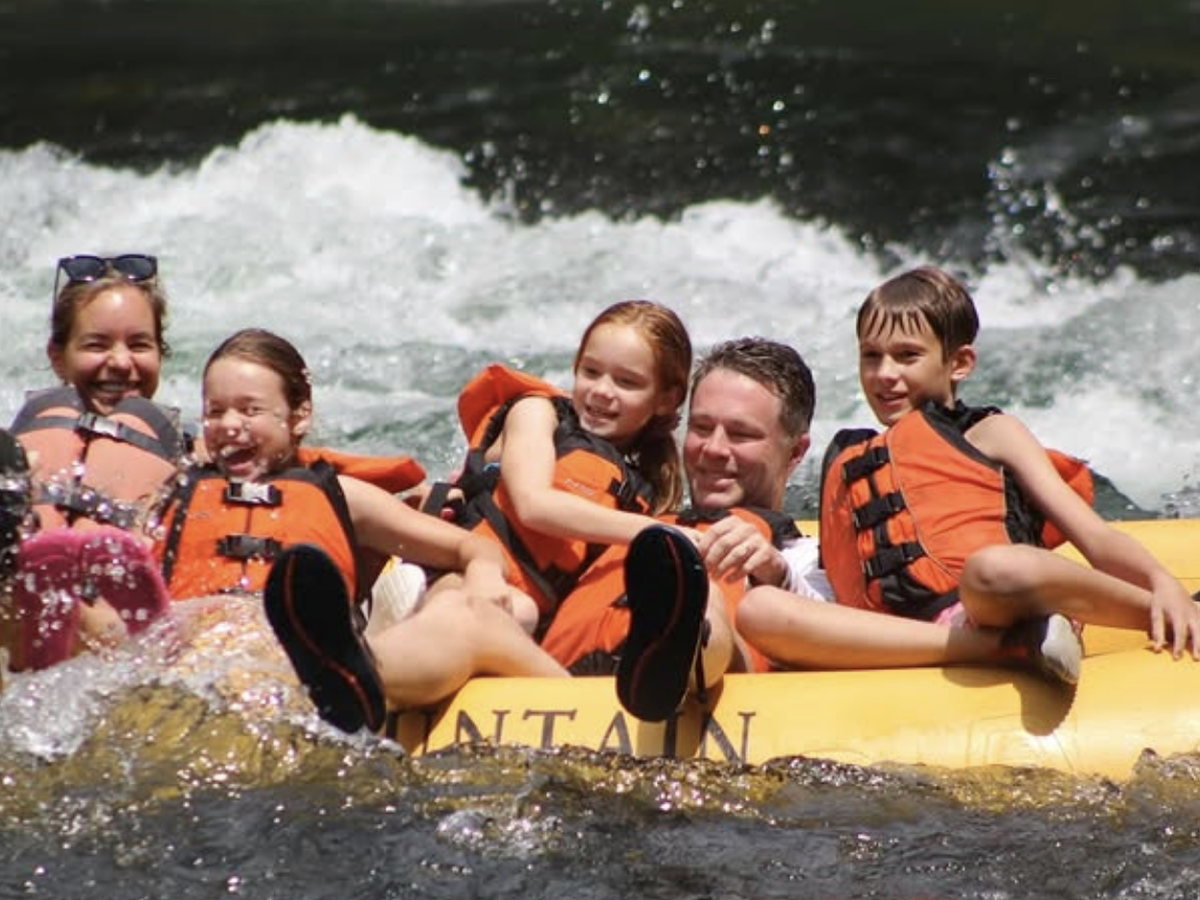 A smiling family in life jackets enjoys a whitewater rafting adventure, splashing through a lively river current on a bright sunny day.