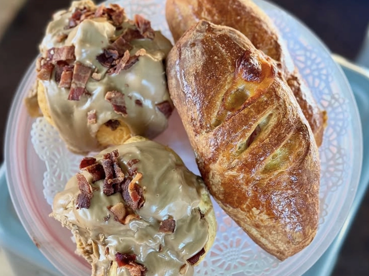 A plate with two maple bacon cinnamon rolls and two crusty golden baguettes sits atop a lacy doily on a round tray.