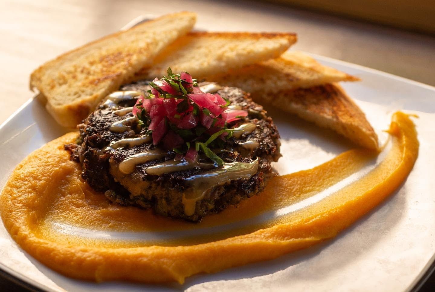 A seared black bean burger patty drizzled with creamy sauce and topped with pickled red onions and fresh herbs sits atop a swirl of golden squash purée. Slices of grilled toast frame the plate, warmly lit by natural sunlight. 