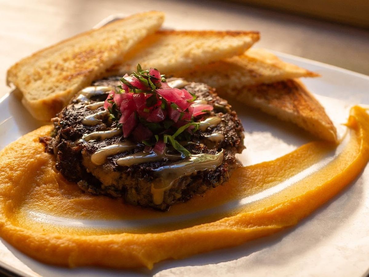 A seared black bean burger patty drizzled with creamy sauce and topped with pickled red onions and fresh herbs sits atop a swirl of golden squash purée. Slices of grilled toast frame the plate, warmly lit by natural sunlight.