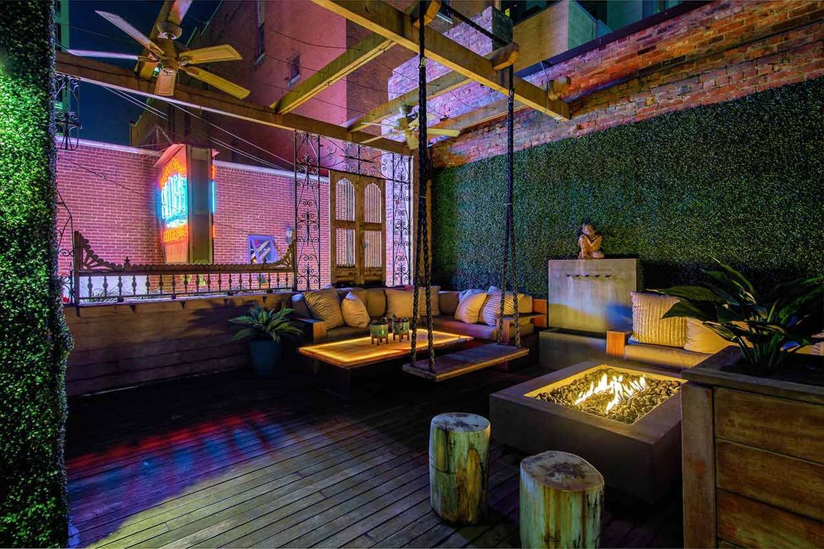 A moody, urban courtyard lounge with hanging swing seating, cushioned banquettes, a modern fire pit, lush green walls, and exposed brick illuminated by warm accent lighting and a neon glow in the background.