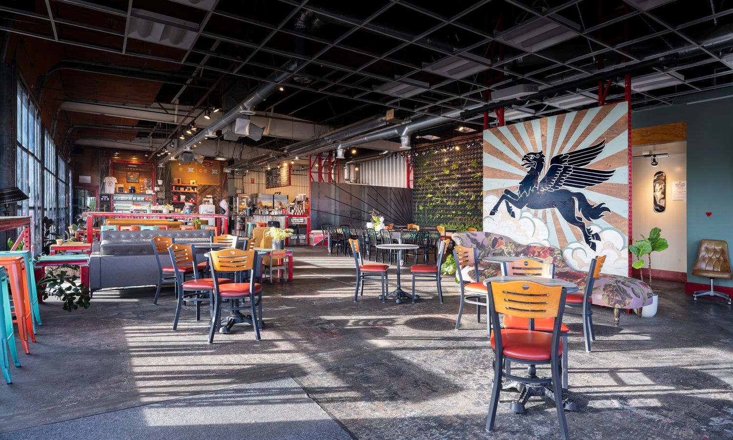 The spacious industrial interior of Iconik Coffee Roasters features exposed ductwork, polished concrete floors, and colorful mismatched café seating in orange, teal, and wood. A bold Pegasus mural with sunburst rays anchors the back wall beside a lush living plant wall. 