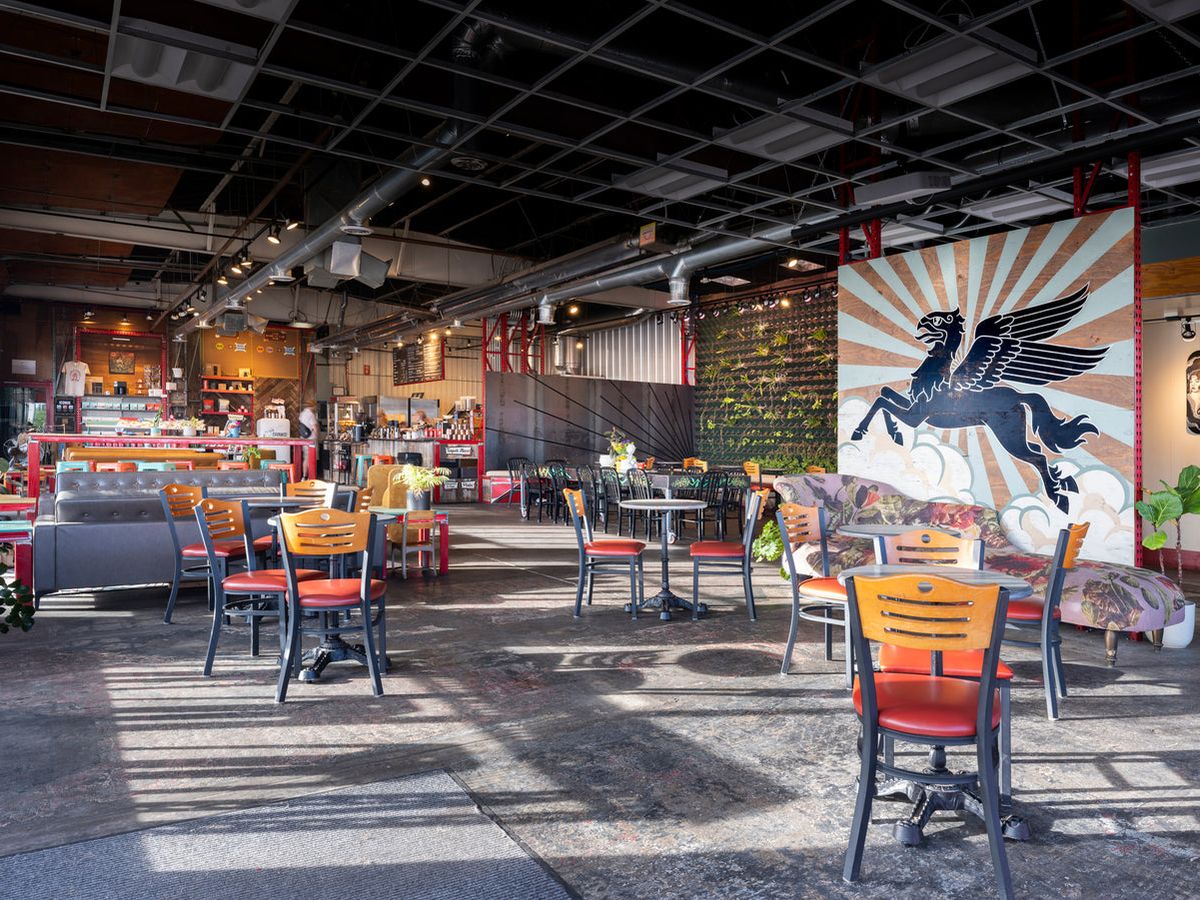 The spacious industrial interior of Iconik Coffee Roasters features exposed ductwork, polished concrete floors, and colorful mismatched café seating in orange, teal, and wood. A bold Pegasus mural with sunburst rays anchors the back wall beside a lush living plant wall.