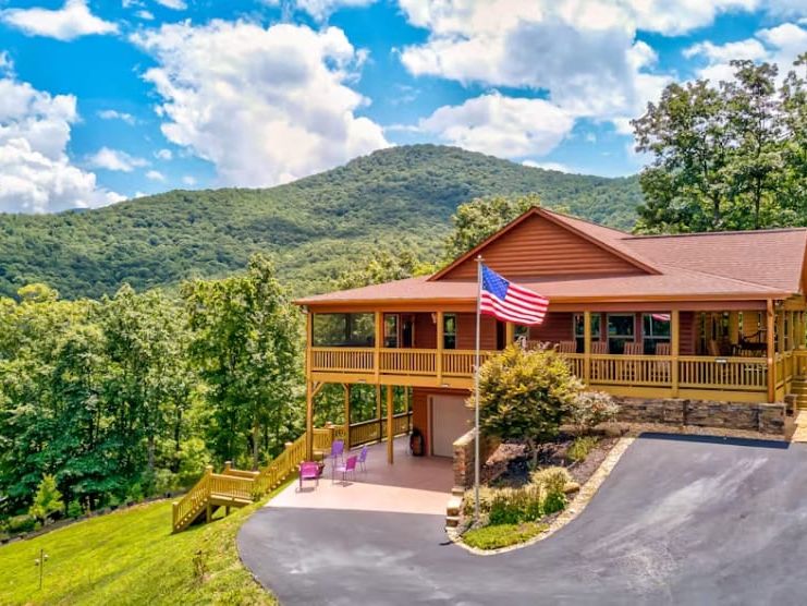 night Sky cabin sits perched on a hilltop with a sweeping wraparound deck flying an American flag, backed by a dramatic view of lush green mountains under a bright blue sky with billowing clouds. The warm wood exterior and open patio below give the property an inviting, patriotic mountain retreat feel. Photo Credit: @suchesandblairsvillesvrproperties
