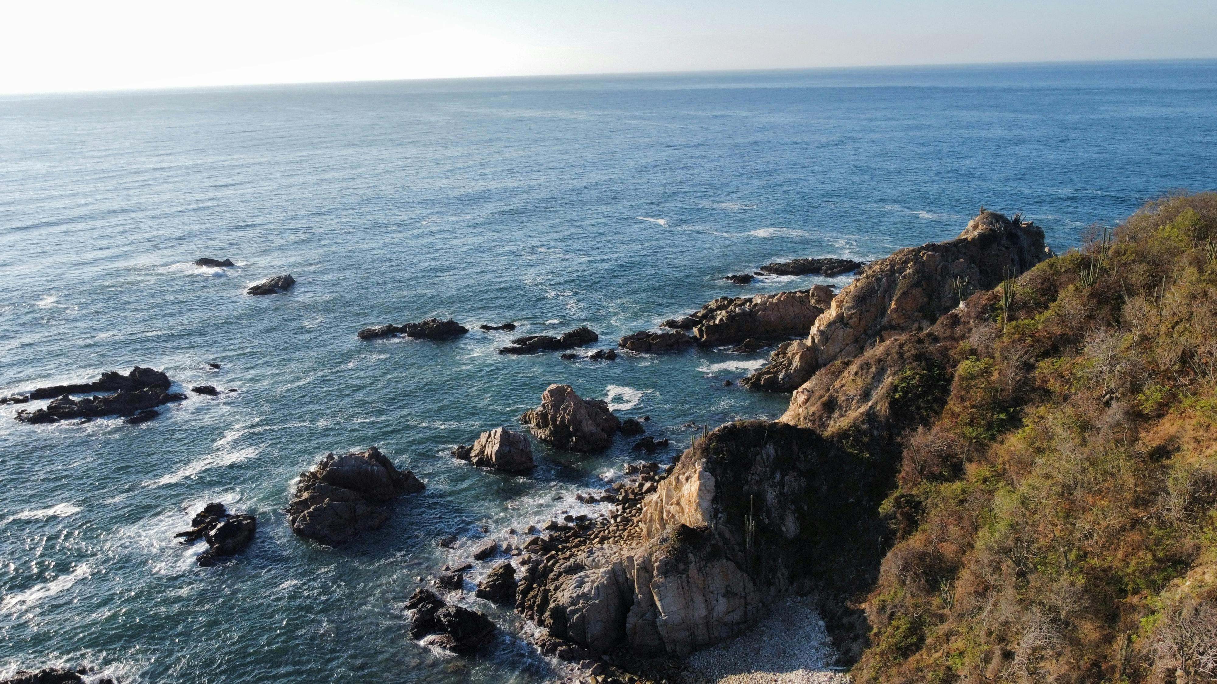Rocky coastal cliffs overlook the deep blue ocean with waves crashing against the shore. The rugged landscape and clear horizon create a peaceful seaside view. This scenic coastline highlights the natural beauty of Mexico’s Pacific coast.