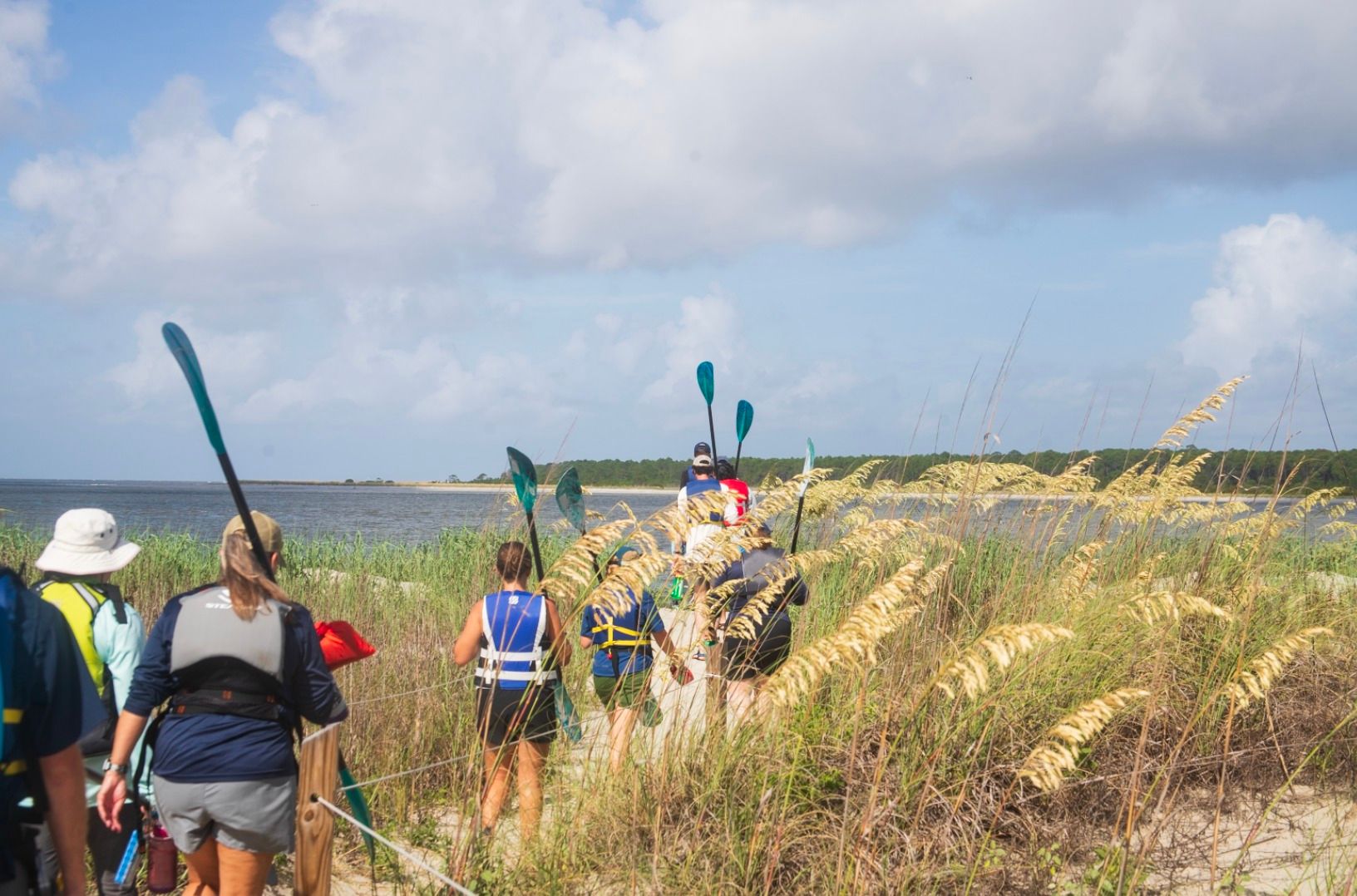 A small group of paddlers walks through tall coastal grass toward the water, carrying their kayaks and gear. The sky is bright and the calm shoreline stretches ahead, setting the scene for a fun day outdoors. It captures the simple joy of exploring Kiawah by paddle.