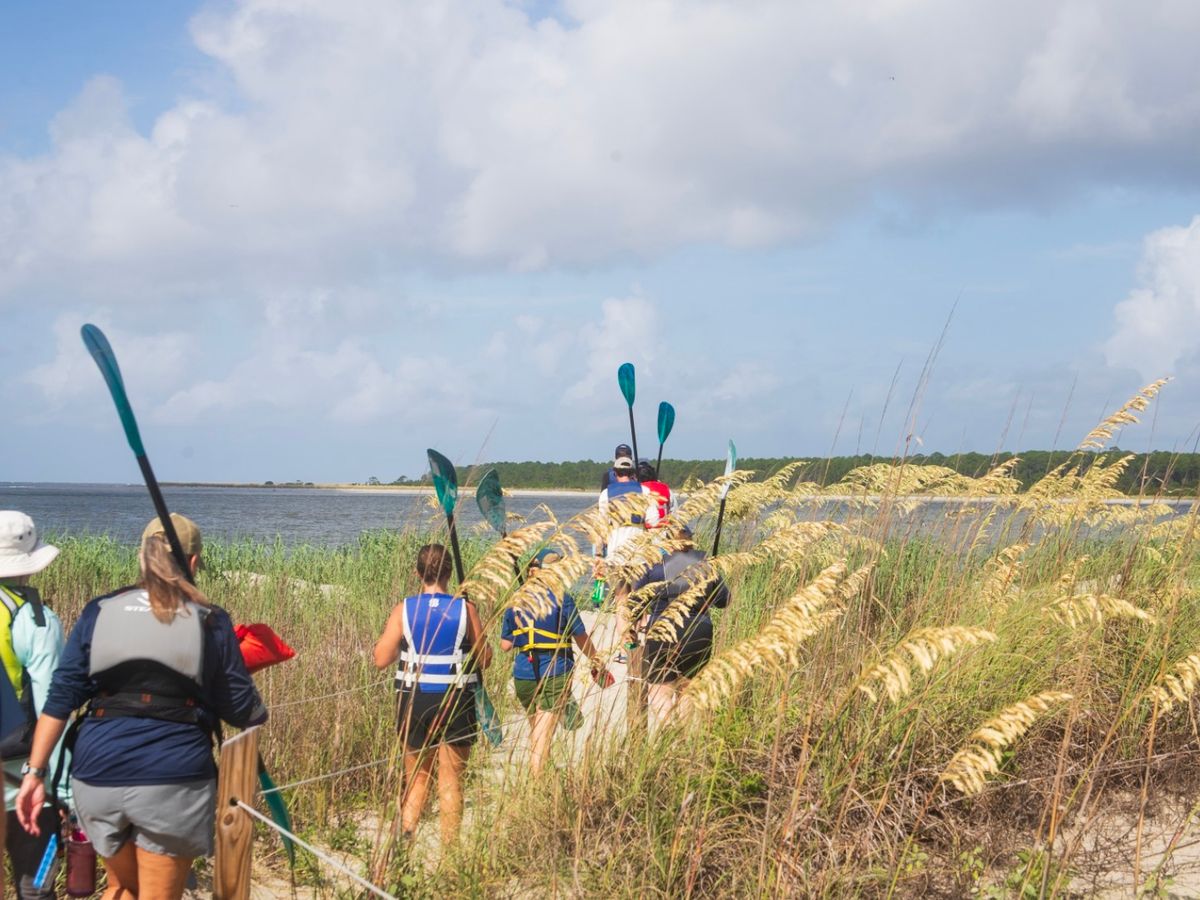 A small group of paddlers walks through tall coastal grass toward the water, carrying their kayaks and gear. The sky is bright and the calm shoreline stretches ahead, setting the scene for a fun day outdoors. It captures the simple joy of exploring Kiawah by paddle.