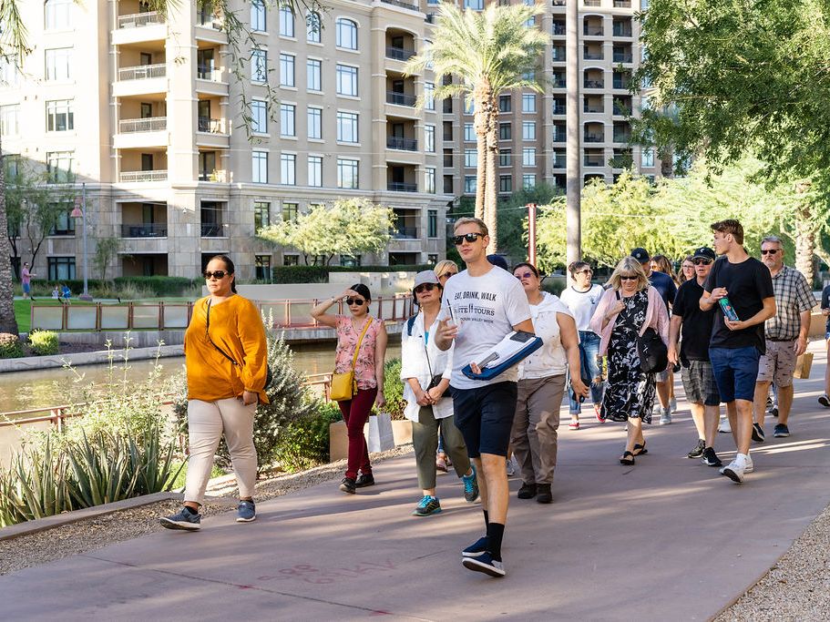 A lively group strolls along a sunny canal path, enjoying a guided food tour through the city. The mix of modern architecture and palm trees creates the perfect urban backdrop for tasting and exploring.