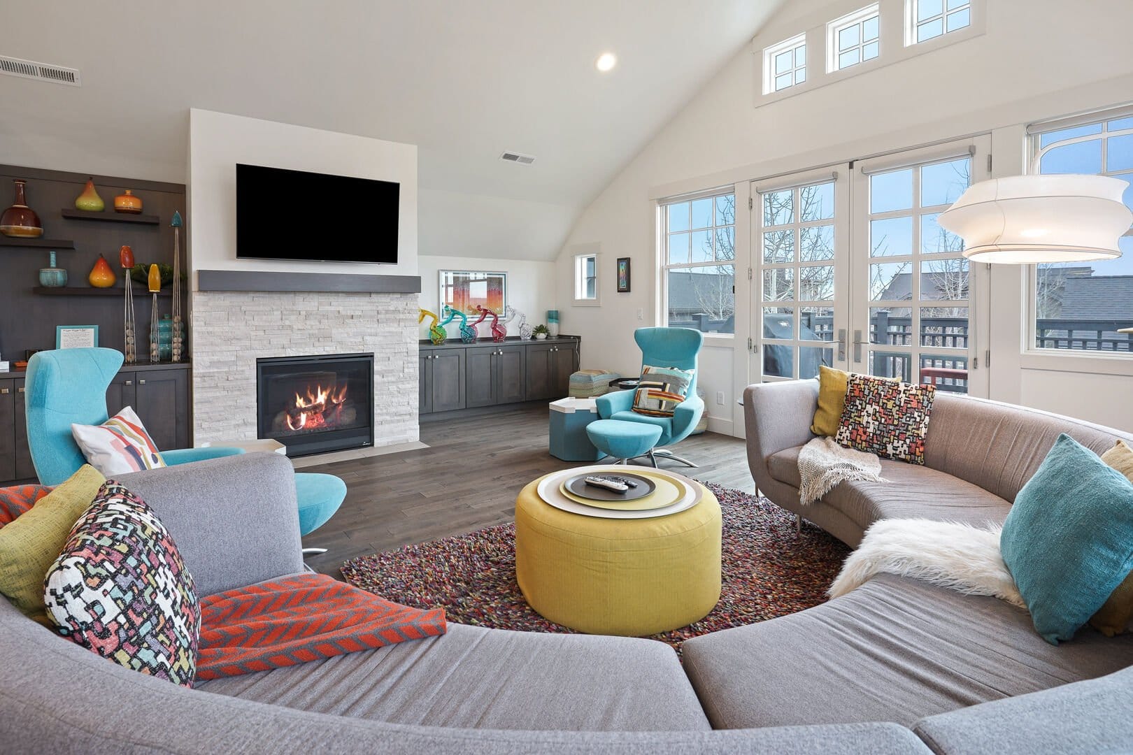 The vibrant living room at Cairn vacation rental features a curved gray sectional sofa and bold teal accent chairs arranged around a yellow ottoman, with a lit stone fireplace and mounted TV as the focal point. Vaulted ceilings, colorful artwork, and large French doors leading to a balcony give the space an energetic, playful character.