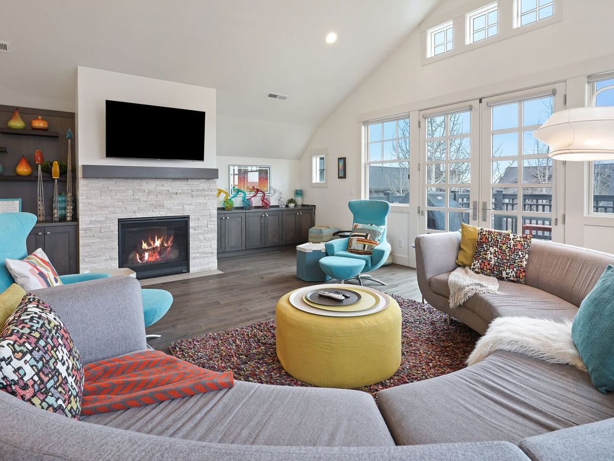 The vibrant living room at Cairn vacation rental features a curved gray sectional sofa and bold teal accent chairs arranged around a yellow ottoman, with a lit stone fireplace and mounted TV as the focal point. Vaulted ceilings, colorful artwork, and large French doors leading to a balcony give the space an energetic, playful character.