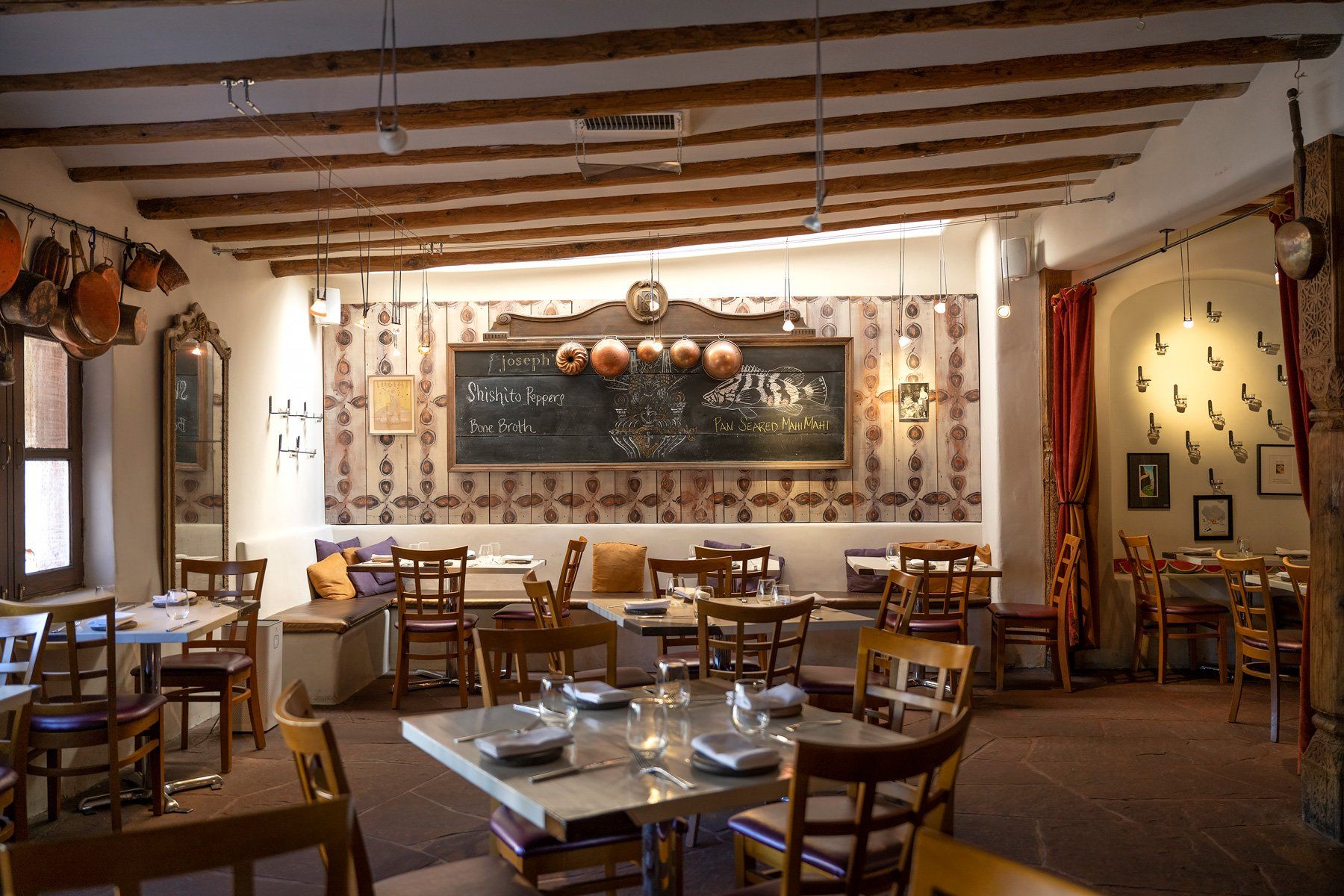A cozy dining room with exposed wooden beams, adobe walls, and terracotta floors centers on a large chalkboard menu listing shishito peppers, bone broth, and pan-seared mahi mahi. Copper pots hang from the wall and pendant lights cast a warm glow over the simply set tables.