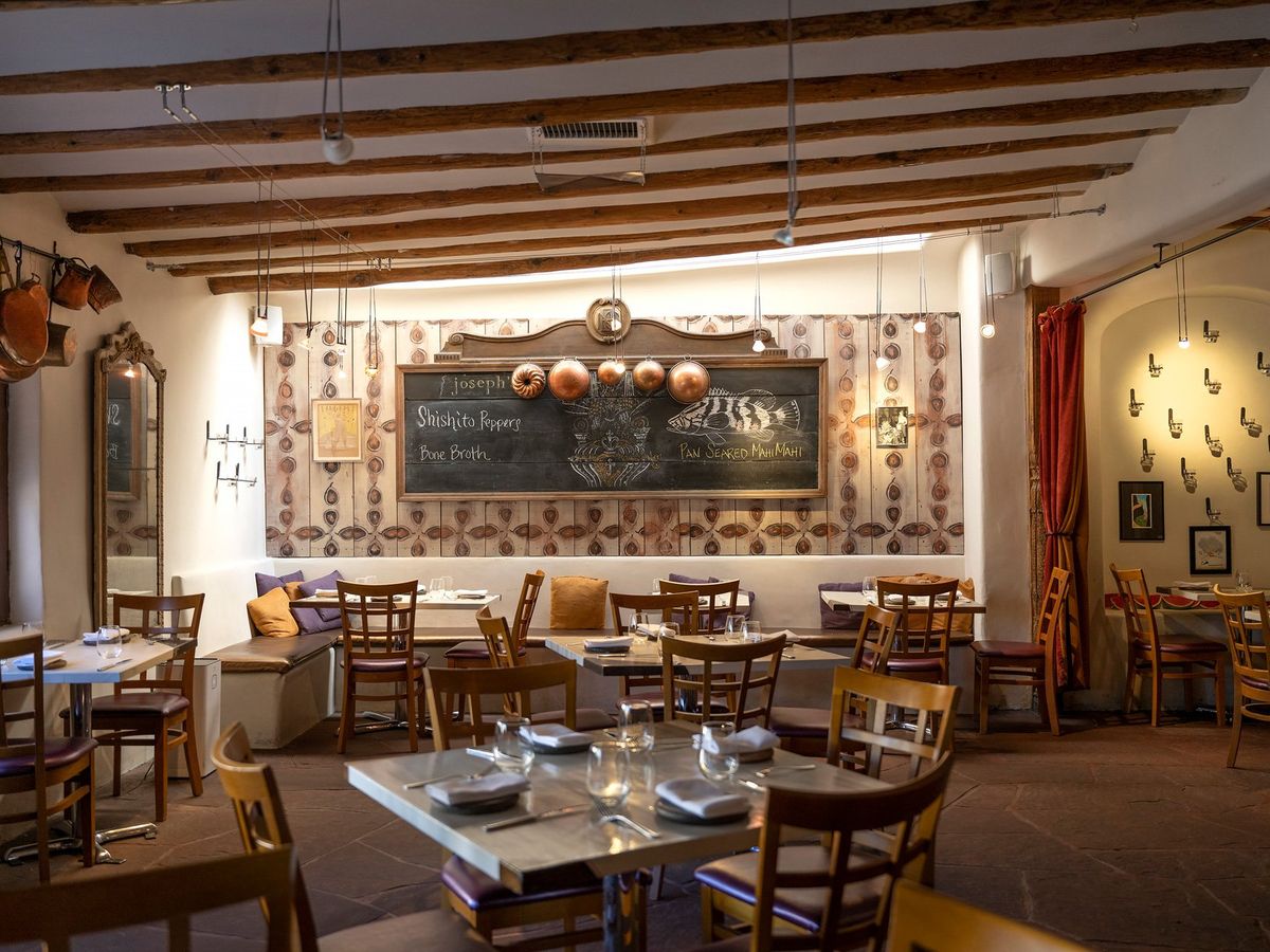 A cozy dining room with exposed wooden beams, adobe walls, and terracotta floors centers on a large chalkboard menu listing shishito peppers, bone broth, and pan-seared mahi mahi. Copper pots hang from the wall and pendant lights cast a warm glow over the simply set tables.