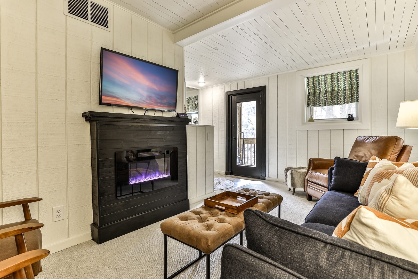 Cozy living room with a black fireplace and mounted TV, leather armchair, modern sofa with patterned pillows, and a tufted ottoman table.
