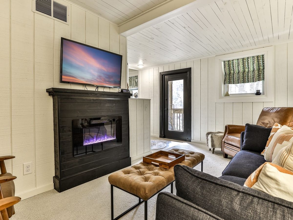 Cozy living room with a black fireplace and mounted TV, leather armchair, modern sofa with patterned pillows, and a tufted ottoman table.
