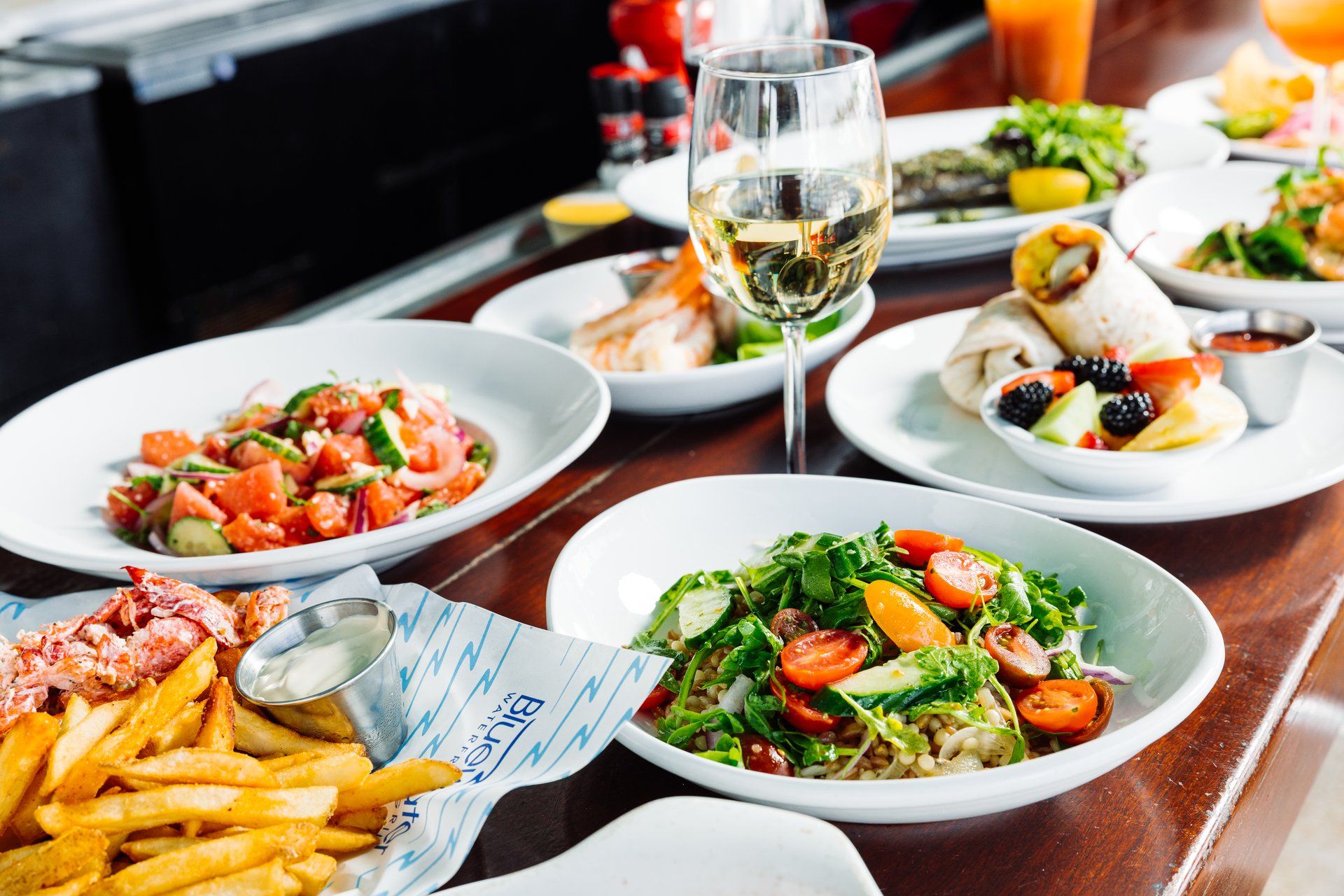 A colorful spread of fresh seafood, salads, and fries paired with a chilled glass of white wine at Bluewater Waterfront Grill. The bright dishes and casual coastal setting capture the relaxed dining vibe by the marina.