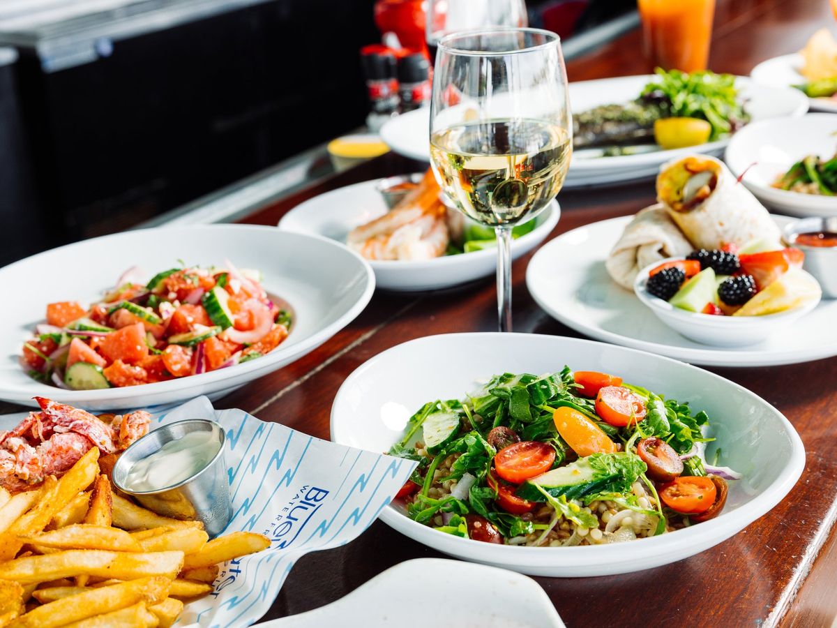 A colorful spread of fresh seafood, salads, and fries paired with a chilled glass of white wine at Bluewater Waterfront Grill. The bright dishes and casual coastal setting capture the relaxed dining vibe by the marina.