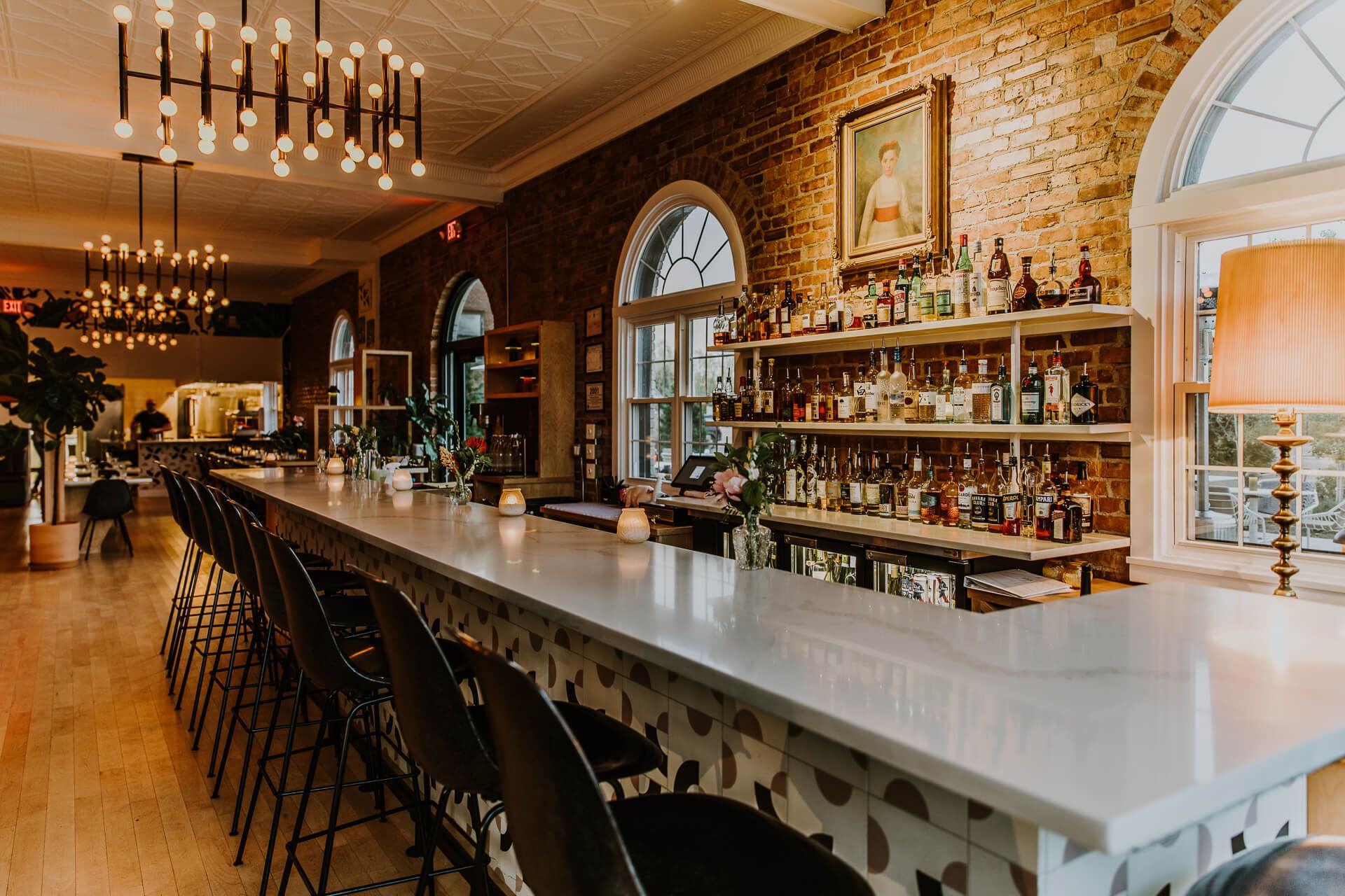 A beautifully restored bar stretches down a warmly lit dining room with exposed brick walls, arched leaded-glass windows, and a marble countertop lined with black stools. Shelves of liquor bottles and a vintage portrait complete the elegant, historic ambiance.
