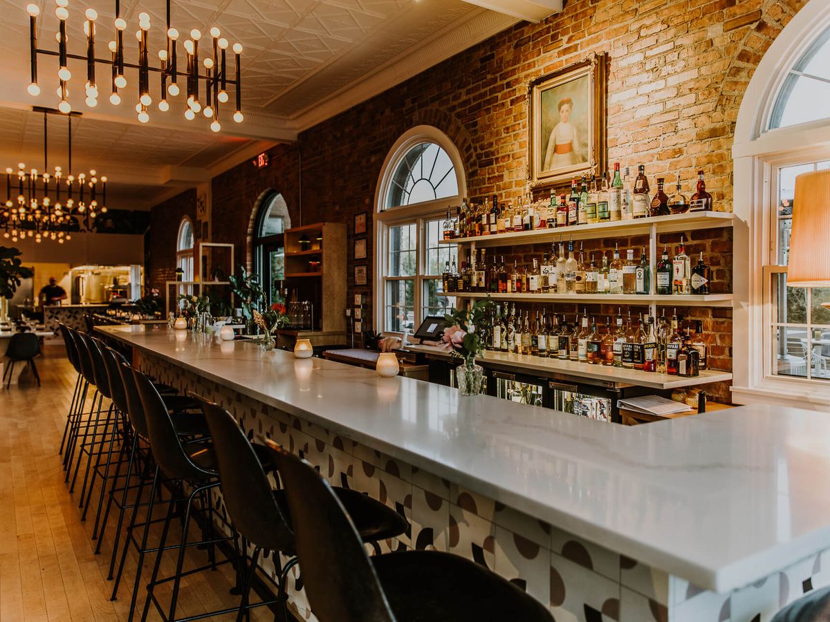 A beautifully restored bar stretches down a warmly lit dining room with exposed brick walls, arched leaded-glass windows, and a marble countertop lined with black stools. Shelves of liquor bottles and a vintage portrait complete the elegant, historic ambiance.