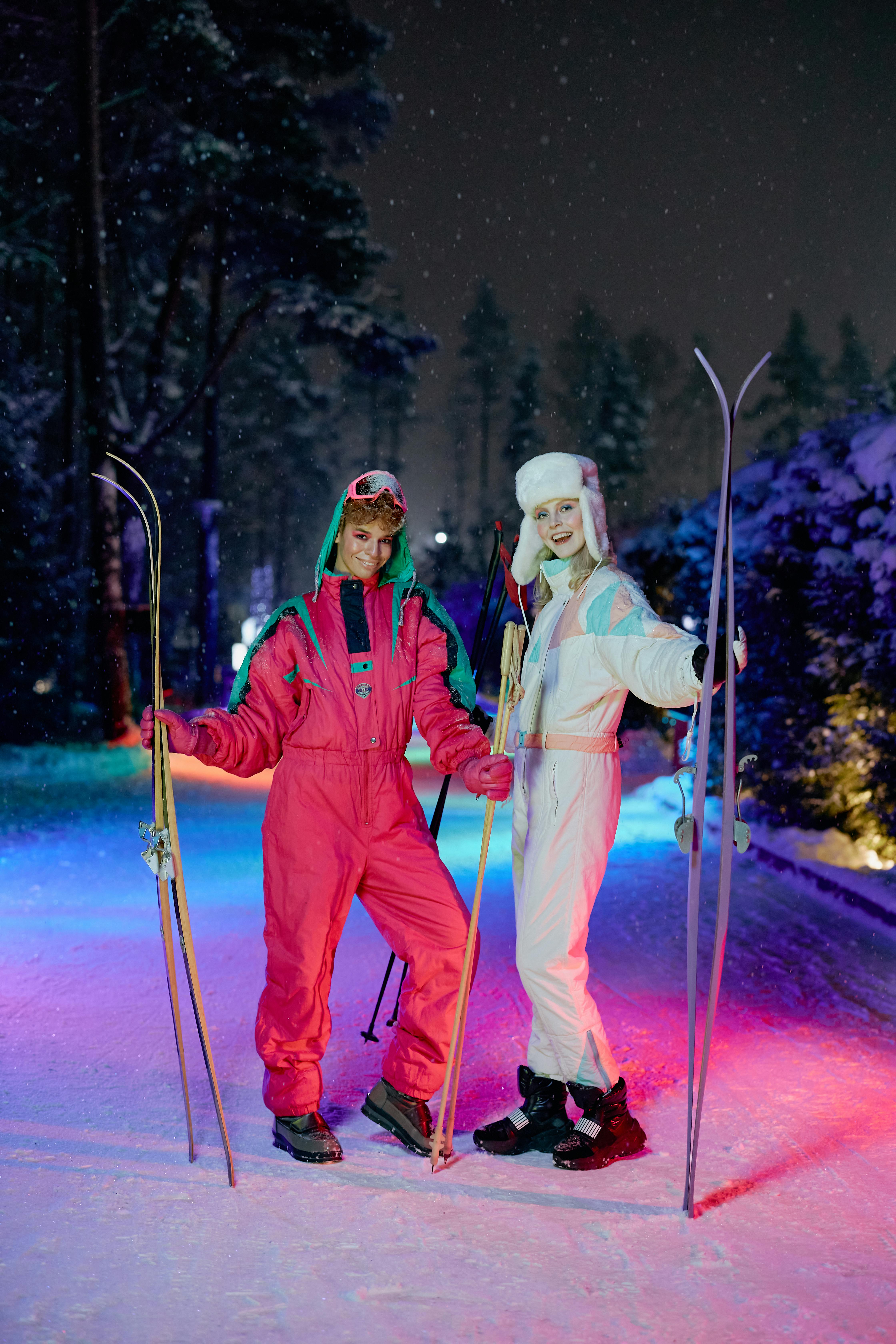 Two women in bright retro ski suits pose with their skis on a snowy trail at night. Snow falls gently around them as colorful lights reflect off the snow, creating a fun and vibrant winter ski scene. Their vintage ski outfits and smiles capture the joy of a cold evening on the slopes.