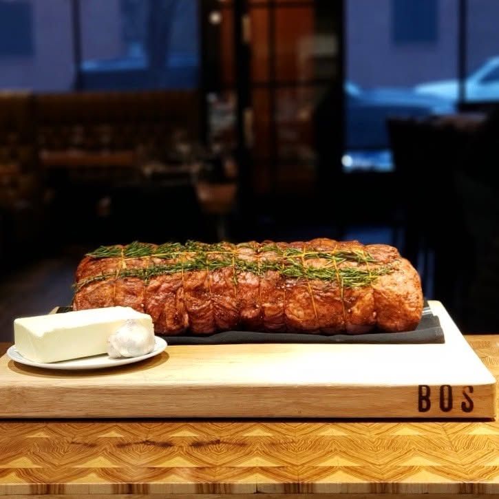 A large seared roast tied with fresh rosemary sprigs rests on a slate slab atop a thick butcher-block cutting board branded "BOS." A plate holding a block of butter and a bulb of garlic sits alongside in the warmly lit, upscale steakhouse setting.
