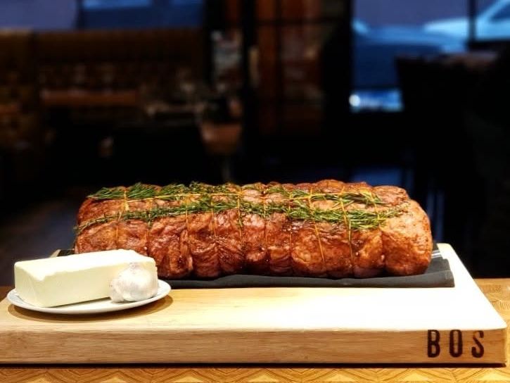 A large seared roast tied with fresh rosemary sprigs rests on a slate slab atop a thick butcher-block cutting board branded "BOS." A plate holding a block of butter and a bulb of garlic sits alongside in the warmly lit, upscale steakhouse setting.