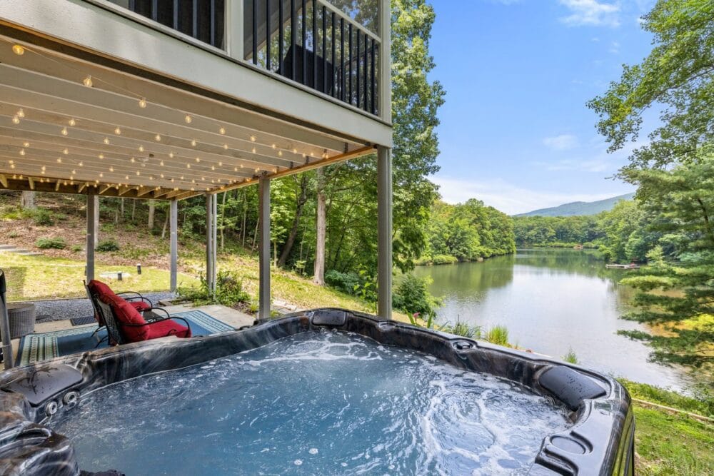A bubbling hot tub on a patio overlooking a serene lake surrounded by lush forest, with string lights hanging from the deck above and mountains visible in the distance.