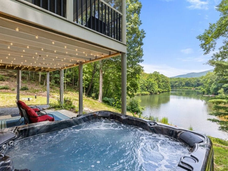 A bubbling hot tub on a patio overlooking a serene lake surrounded by lush forest, with string lights hanging from the deck above and mountains visible in the distance.