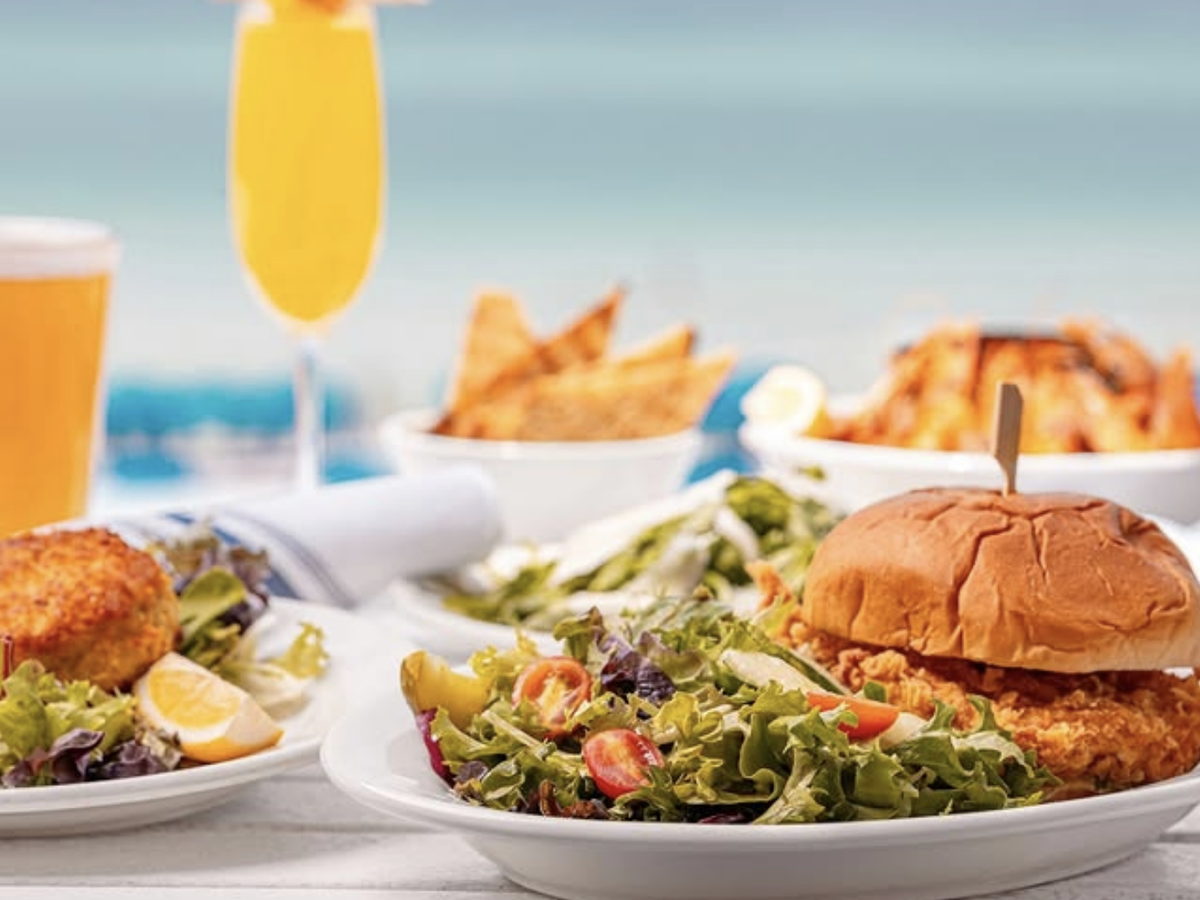 Beachfront dining table with a crispy chicken sandwich, fresh side salad, crab cake, tortilla chips, and cold drinks including beer and a mimosa.