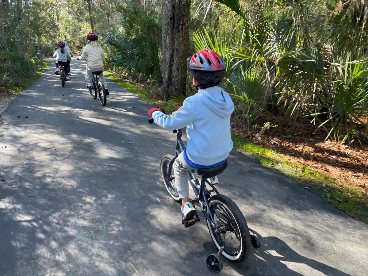 Kids riding bikes along a shaded paved trail in Charleston County Parks. These family-friendly bike paths are perfect for easy rides surrounded by trees and nature.