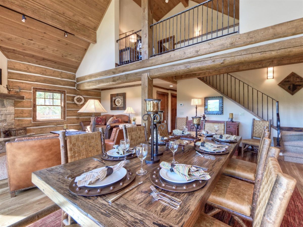 A rustic dining and living area in a log cabin features a wooden table set for eight, leather chairs, exposed beams, and a lofted second floor with iron railings.