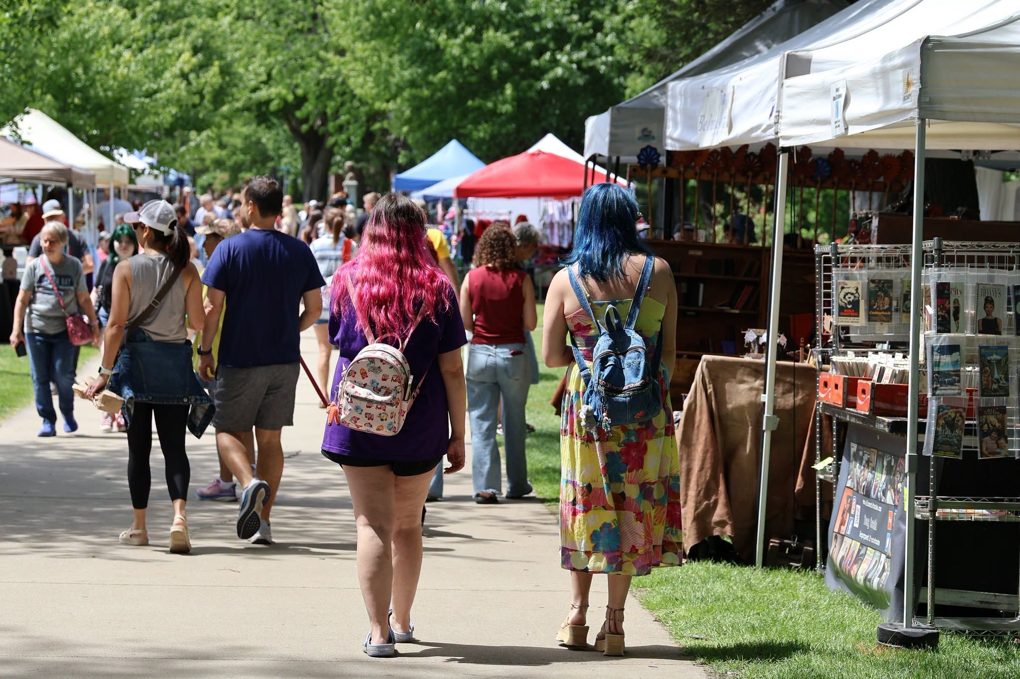  Crowds of shoppers stroll along a sunny, tree-lined path browsing white vendor tents at an outdoor antique and flea market. Two women with brightly colored pink and blue hair walk together in the foreground past a booth of vintage books and collectibles.