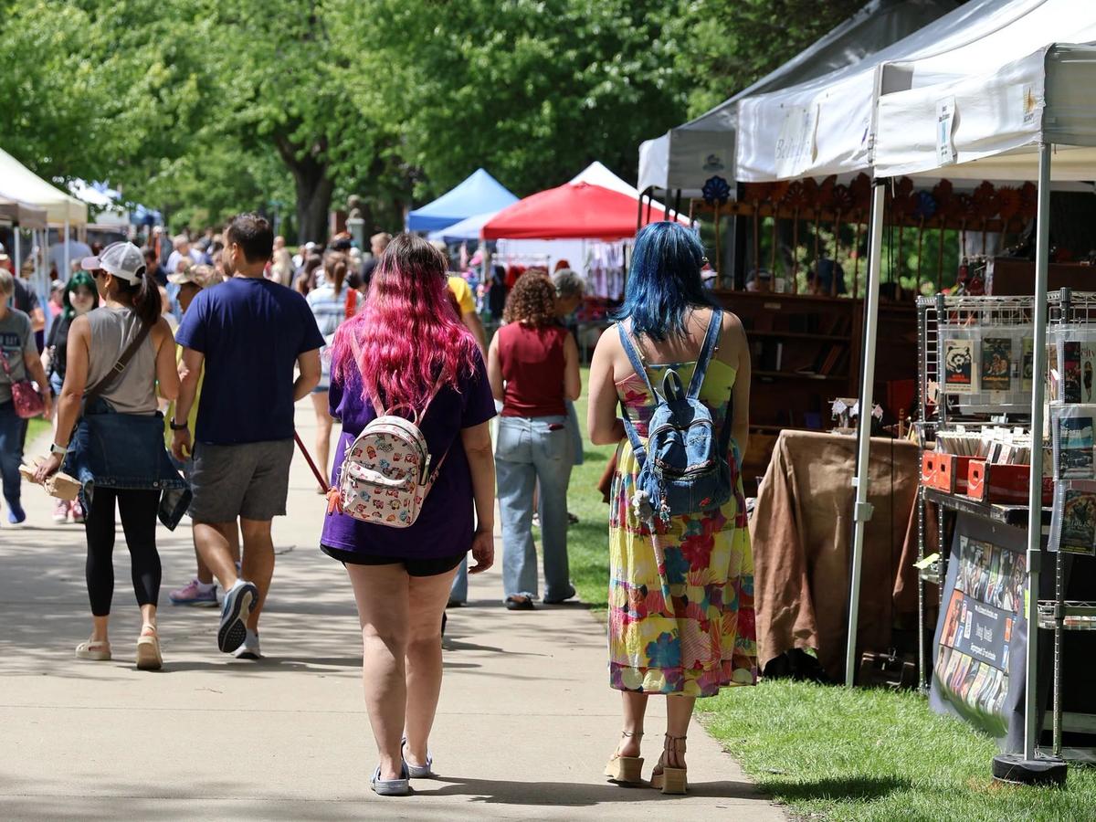 Crowds of shoppers stroll along a sunny, tree-lined path browsing white vendor tents at an outdoor antique and flea market. Two women with brightly colored pink and blue hair walk together in the foreground past a booth of vintage books and collectibles.