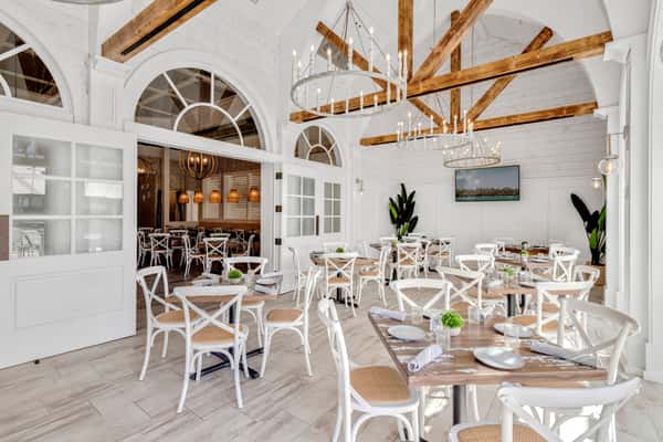 A bright, coastal-inspired restaurant featuring whitewashed walls, wooden beams, and elegant chandeliers. The airy design with soft natural light creates a relaxed yet stylish dining atmosphere perfect for brunch or evening cocktails.