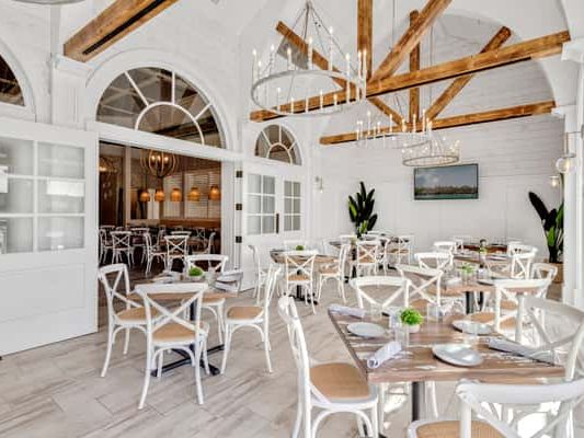 A bright, coastal-inspired restaurant featuring whitewashed walls, wooden beams, and elegant chandeliers. The airy design with soft natural light creates a relaxed yet stylish dining atmosphere perfect for brunch or evening cocktails.