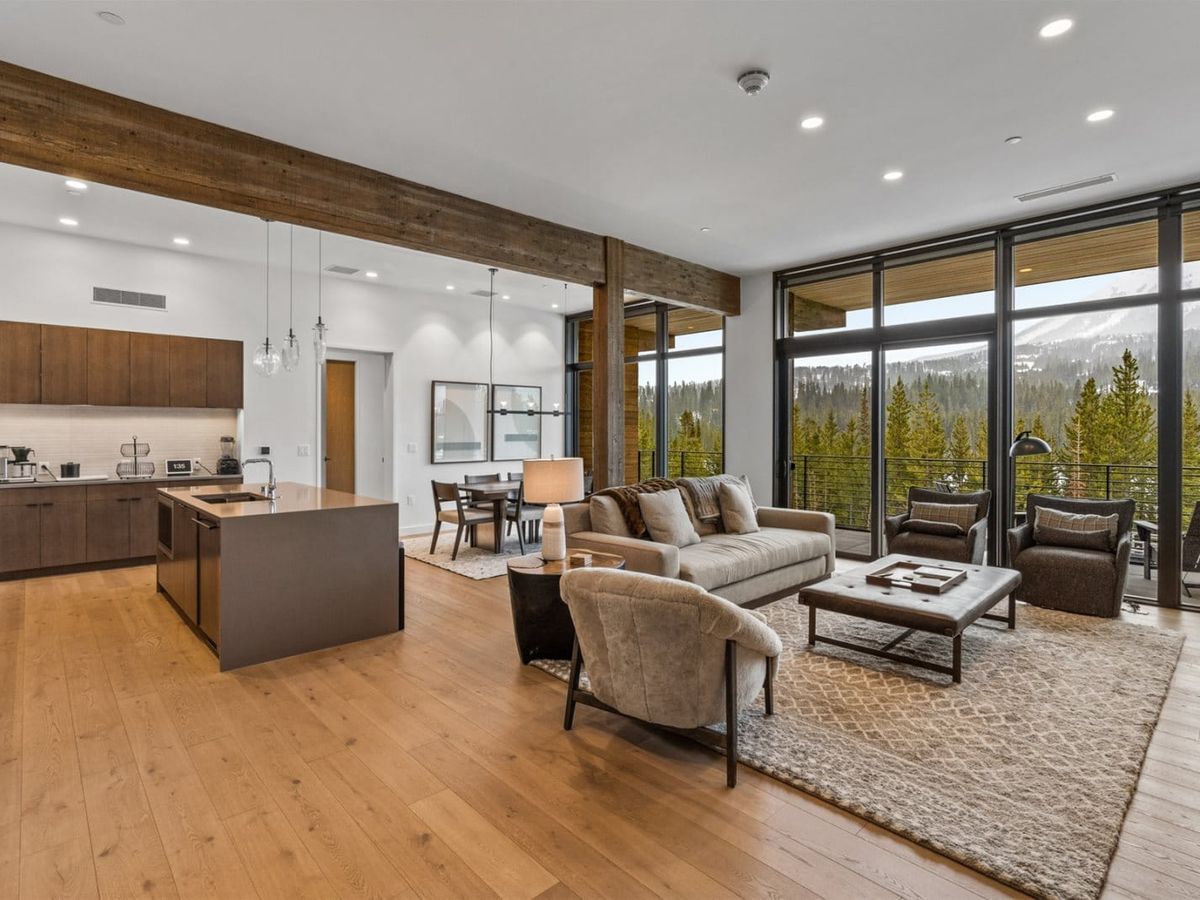 A bright and modern living room with floor-to-ceiling windows opens to stunning forest and mountain views. The open kitchen and cozy furniture make this space ideal for relaxing after a day on the slopes.