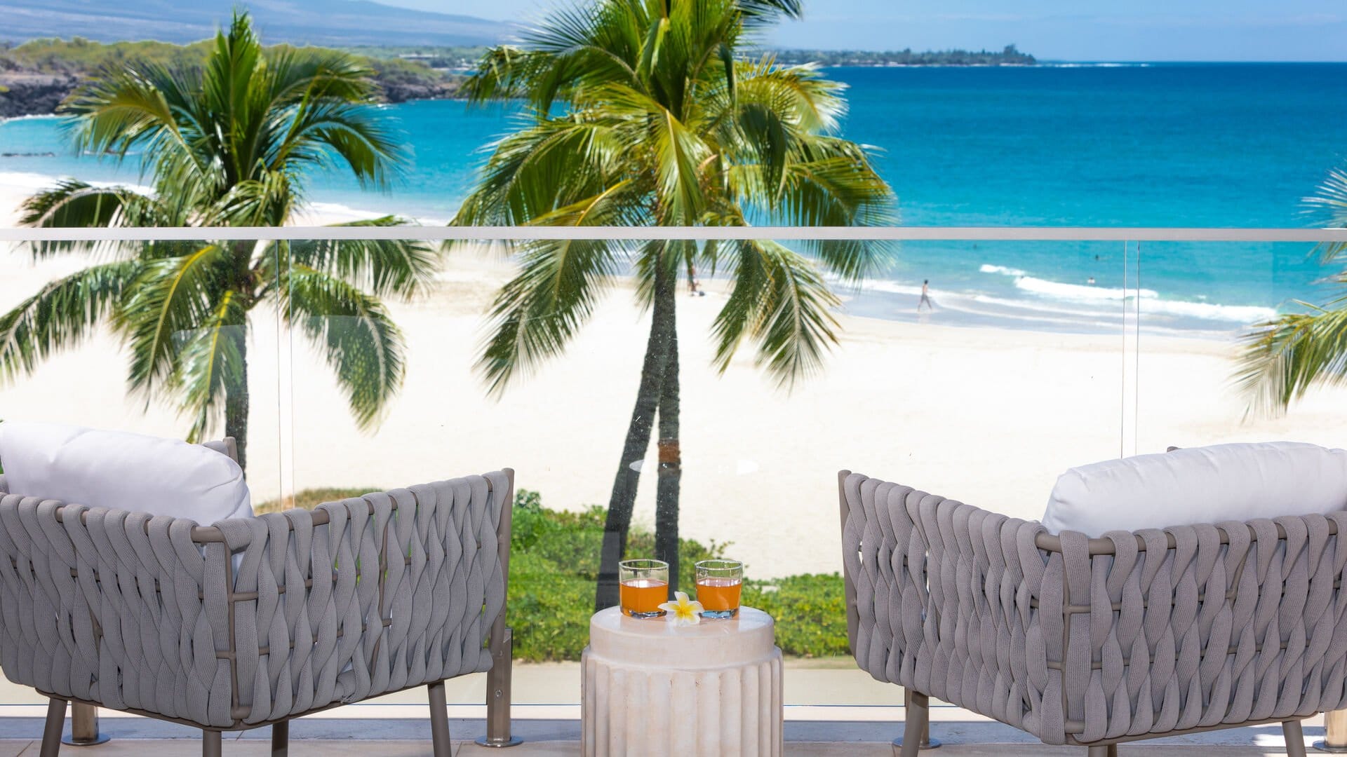 Two cushioned chairs face a turquoise ocean and white sandy beach, separated by palm trees swaying in the breeze, with a small table holding drinks and a plumeria flower between them.