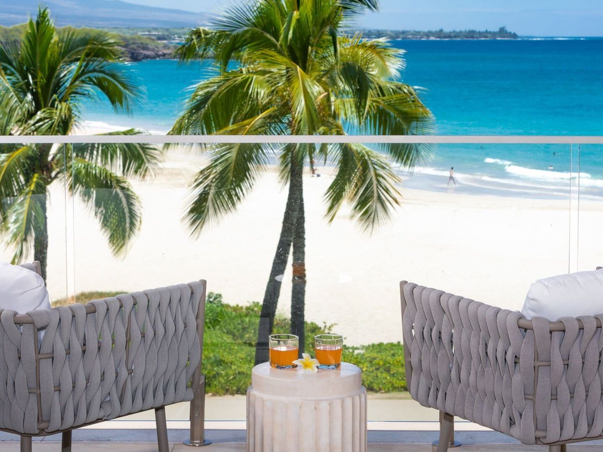 Two cushioned chairs face a turquoise ocean and white sandy beach, separated by palm trees swaying in the breeze, with a small table holding drinks and a plumeria flower between them.